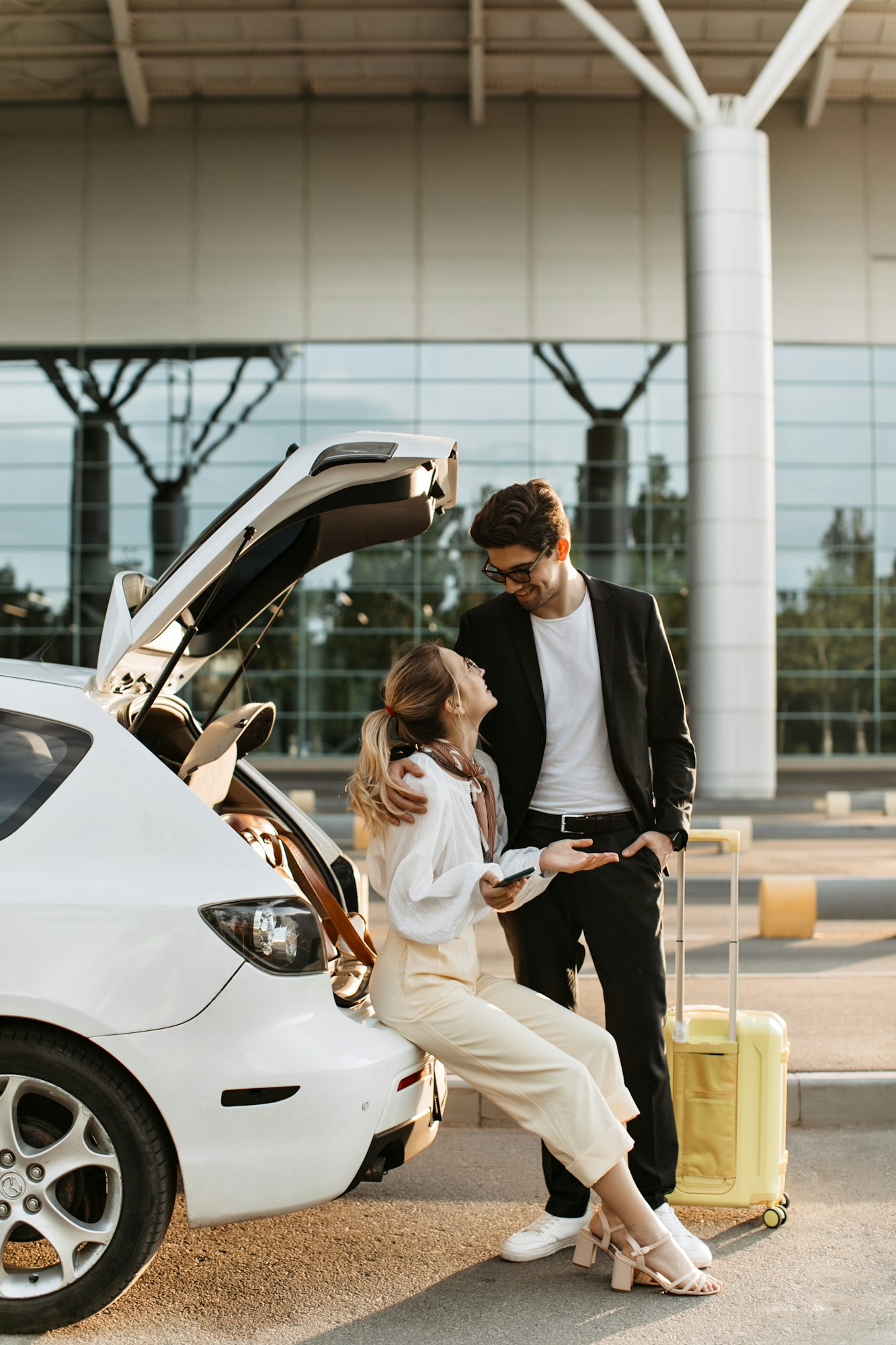 Blonde woman in white outfit looks surprised and sits in car near airport. Brunette man hugs girlfr